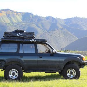 An SUV car with side awning is parked on a field surrounded by mountains.