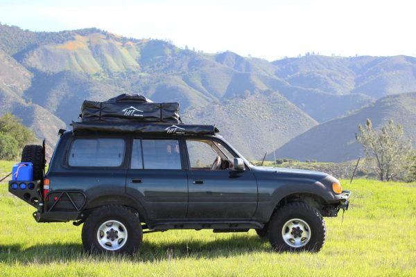 An SUV car with side awning is parked on a field surrounded by mountains.